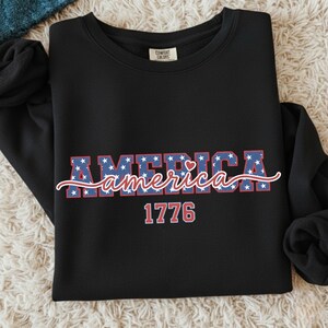 May include: Black sweatshirt with the word "AMERICA" in blue letters filled with white stars. The word "america" is written in a cursive font, with the year "1776" below it in red.