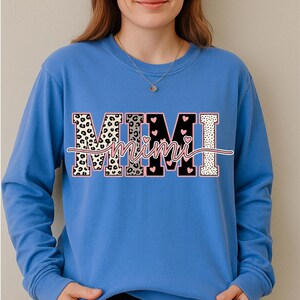 May include: A blue long-sleeve sweatshirt featuring the word "MIMI" in a decorative design. The letters have a leopard print, black with pink hearts, and white polka dot pattern. The word "Mimi" is in a cursive font.