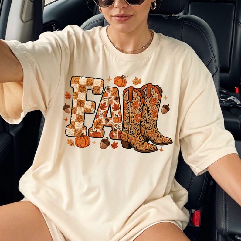 K&ouml;nnte beinhalten: Cremefarbenes T-Shirt mit dem Wort "FALL" in einem herbstlichen Design. Das "F" ist kariert, das "A" ist mit Bl&auml;ttern gef&uuml;llt und das "LL" wird durch ein Paar Cowboystiefel ersetzt. K&uuml;rbisse, Eicheln und Bl&auml;tter umgeben den Text.