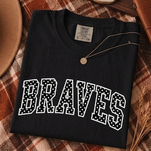 Op de afbeelding: Een zwart T-shirt met korte mouwen en een ronde hals, voorzien van het woord BRAVES in grote, witte blokletters met een zwart stippenpatroon. Het shirt heeft een Comfort Colors merklabel in de nek en een label met maat S.