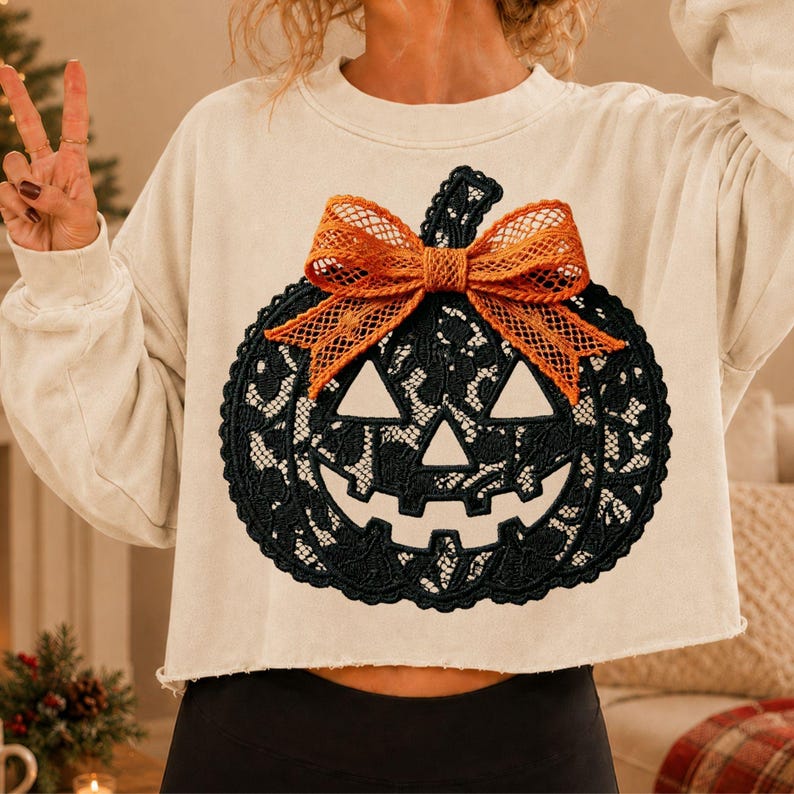 May include: A cream-colored long-sleeve sweatshirt featuring a large black lace pumpkin graphic on the front. The pumpkin design includes a prominent orange textured bow tied at the top stem. The sweatshirt has a cropped hem and long sleeves.