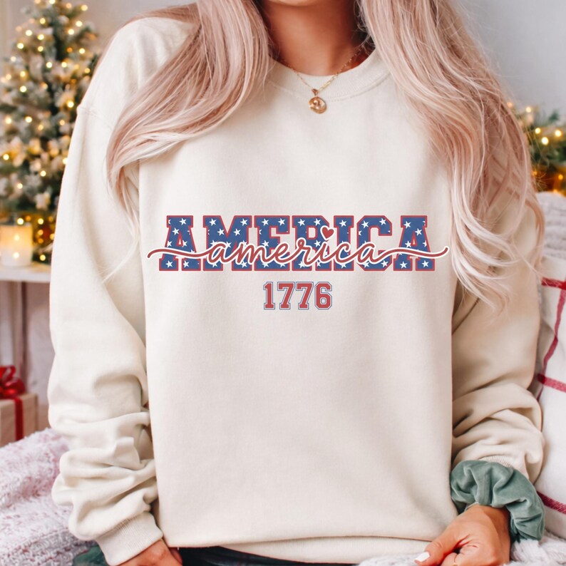 May include: Cream-colored sweatshirt with the word "AMERICA" in blue, star-filled letters, and the word "America" in a cursive font. The number "1776" is below the word America. The sweatshirt is a casual style.