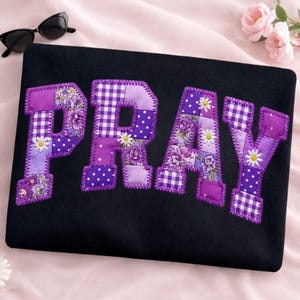 Könnte beinhalten: Schwarzes Stoffstück mit dem Wort "PRAY" in lila Patchwork-Buchstaben appliziert. Die Buchstaben weisen Blumen-, Tupfen- und Vichy-Muster auf. Sonnenbrille und Blumen sind im Hintergrund.