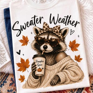 May include: A white t-shirt featuring a graphic of a raccoon wearing a leopard print headband and sunglasses, holding a coffee cup labeled Pumpkin Spice. The text Sweater Weather is printed above, surrounded by orange autumn maple leaves.