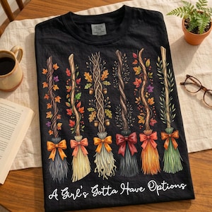 May include: Black t-shirt featuring a design of six decorative brooms with autumn leaves and bows in various colors. The text "A Girl's Gotta Have Options" is printed at the bottom of the shirt.