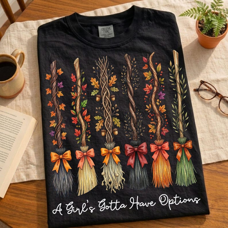 May include: Black t-shirt featuring a design of six decorative brooms with autumn leaves and bows in various colors. The text "A Girl's Gotta Have Options" is printed at the bottom of the shirt.