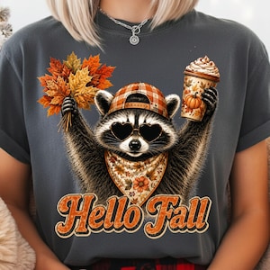 Könnte beinhalten: Ein dunkelgraues T-Shirt mit einer Grafik eines Waschbären, der eine karierte Baseballkappe, herzförmige Sonnenbrillen und ein Blumenbandana trägt. Der Waschbär hält einen Strauß Herbstblätter und einen Kaffeebecher mit Kürbisaufdruck. Text Hello Fall.