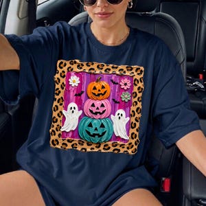 Puede incluir: Una camiseta azul marino con un gr&aacute;fico cuadrado con borde de estampado de leopardo. El dise&ntilde;o incluye una pila de tres calabazas en naranja, rosa y turquesa, dos fantasmas blancos, peque&ntilde;os murci&eacute;lagos negros y tres flores peque&ntilde;as sobre un fondo morado.