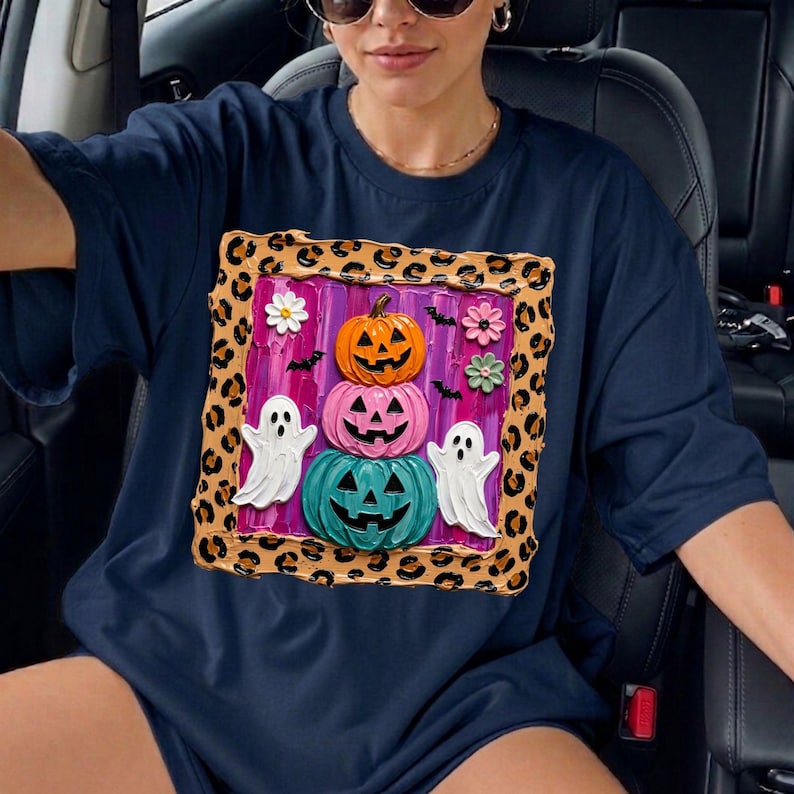 Puede incluir: Una camiseta azul marino con un gr&aacute;fico cuadrado con borde de estampado de leopardo. El dise&ntilde;o incluye una pila de tres calabazas en naranja, rosa y turquesa, dos fantasmas blancos, peque&ntilde;os murci&eacute;lagos negros y tres flores peque&ntilde;as sobre un fondo morado.