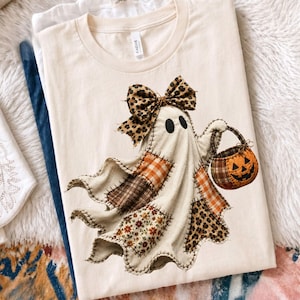 Pu&ograve; includere: Una t-shirt a maniche corte color crema con la grafica di un fantasma composto da vari ritagli di tessuto, inclusi motivi a quadri, floreali e leopardati. Il fantasma indossa un fiocco leopardato e tiene in mano un secchiello a zucca arancione.