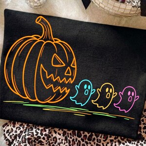 Op de afbeelding: Zwart Halloween-thema grafisch ontwerp met een grote oranje pompoen en drie kleurrijke spookfiguren. Het ontwerp is omlijnd in neonkleuren, wat een spookachtige en feestelijke uitstraling cre&euml;ert. Geschikt voor kleding.