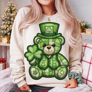 May include: A cream sweatshirt with a green patchwork teddy bear design. The bear wears a top hat and waistcoat, holding a four-leaf clover. The bear is decorated with shamrocks, ideal for St. Patrick's Day celebrations.
