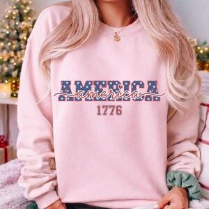 May include: A light pink sweatshirt with the word "AMERICA" in a blue star-patterned font, with a red and white cursive "america" underneath. The number "1776" is printed in red below the word "america".