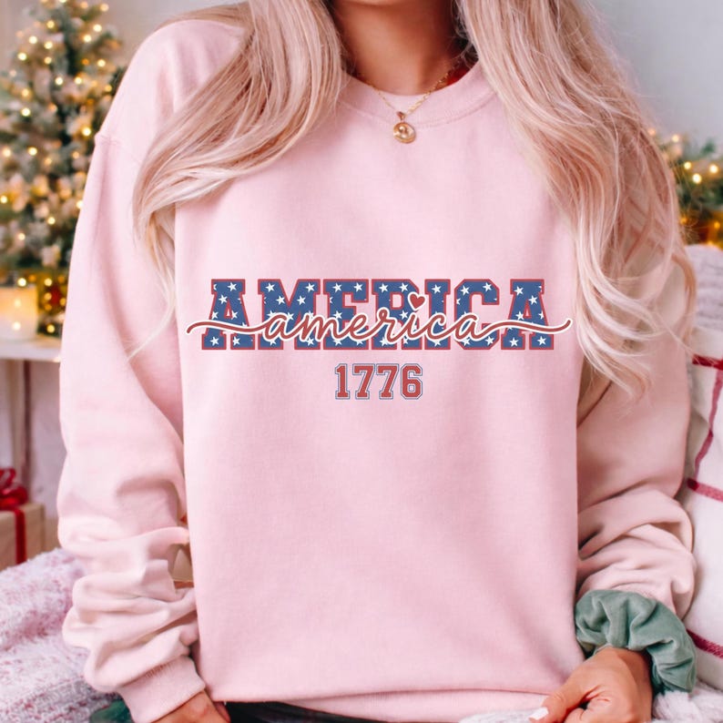 May include: A light pink sweatshirt with the word "AMERICA" in a blue star-patterned font, with a red and white cursive "america" underneath. The number "1776" is printed in red below the word "america".
