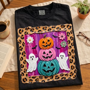 Puede incluir: Una camiseta negra con un gr&aacute;fico cuadrado con borde de estampado de leopardo. El centro muestra una pila de tres calabazas en naranja, rosa y turquesa, flanqueadas por dos fantasmas blancos, peque&ntilde;os murci&eacute;lagos negros y flores rosas y verdes.