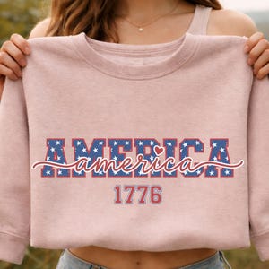 May include: A light pink sweatshirt with the word "AMERICA" in blue letters with white stars, and the word "america" in a cursive font. The number "1776" is below the word "america".