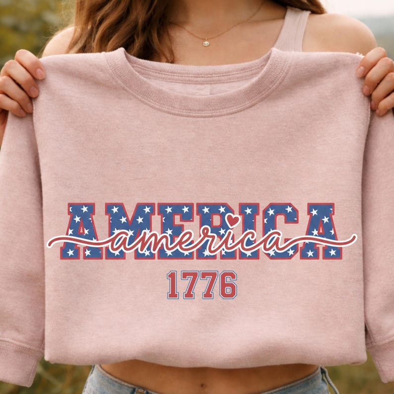 May include: A light pink sweatshirt with the word "AMERICA" in blue letters with white stars, and the word "america" in a cursive font. The number "1776" is below the word "america".