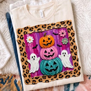 Puede incluir: Una camiseta color crema con un gr&aacute;fico cuadrado con borde de estampado de leopardo. El centro muestra una pila de tres calabazas de Halloween en naranja, rosa y turquesa, flanqueadas por dos fantasmas blancos, murci&eacute;lagos y flores.