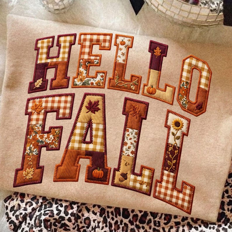 Peut inclure: Un sweat-shirt beige avec le texte HELLO FALL en grandes lettres style bloc. Chaque lettre est remplie d'un patchwork de tissus aux motifs automnaux, incluant du vichy orange et blanc, des motifs floraux et des citrouilles et feuilles brod&eacute;es.