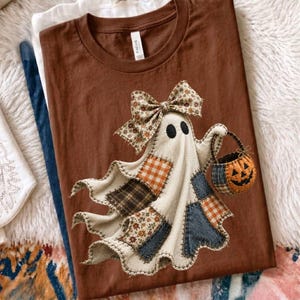Op de afbeelding: Een bruin t-shirt met ronde hals en een afbeelding van een geest die een grote strik met bloemenpatroon op het hoofd draagt. De geest heeft een patchwork-ontwerp met ruitjes- en spijkerstoftexturen en houdt een klein pompoenemmertje vast.