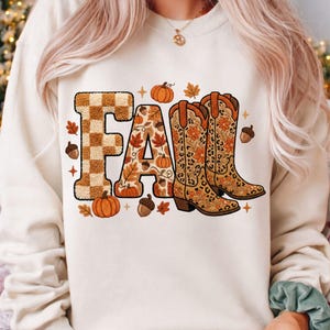 K&ouml;nnte beinhalten: Cremefarbenes Sweatshirt mit einem herbstlichen Motiv. Das Wort "FALL" wird mit einem karierten "F", einem mit Bl&auml;ttern gef&uuml;llten "A" und einem Paar Cowboy-Stiefeln dargestellt. K&uuml;rbisse, Bl&auml;tter und Eicheln umgeben den Text.