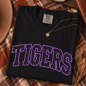Puede incluir: Una camiseta negra de manga corta con la palabra TIGERS en letras grandes y arqueadas de estilo bloque. Las letras son negras con puntos blancos y un contorno morado claro. Un collar dorado descansa sobre el cuello de la camiseta.