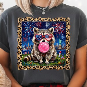 May include: Dark gray t-shirt featuring a raccoon wearing heart-shaped sunglasses with an American flag design, blowing a pink bubble. The raccoon is set against a backdrop of fireworks, the Statue of Liberty, and the New York City skyline.