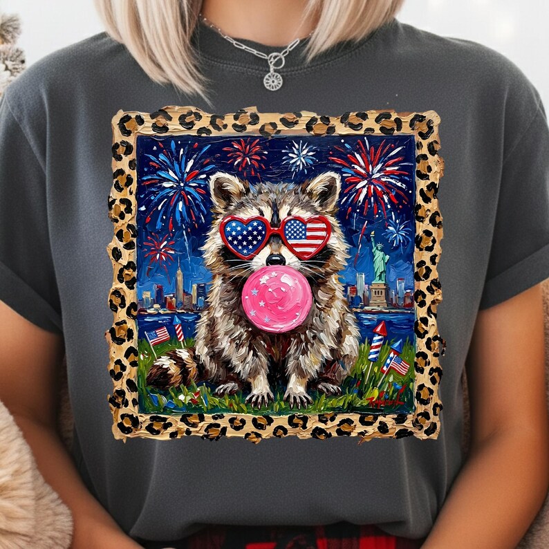 May include: Dark gray t-shirt featuring a raccoon wearing heart-shaped sunglasses with an American flag design, blowing a pink bubble. The raccoon is set against a backdrop of fireworks, the Statue of Liberty, and the New York City skyline.