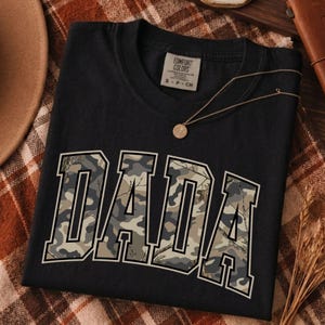Könnte beinhalten: Ein schwarzes Kurzarm-T-Shirt mit dem Wort DADA in großen Blockbuchstaben auf der Brust. Die Buchstaben haben ein Tarnmuster in Grün-, Braun- und Grautönen. Das T-Shirt hat ein Markenetikett von Comfort Colors am Halsausschnitt.