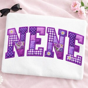 May include: A white fabric surface featuring the word NENE in large, block-style letters. Each letter is constructed from a patchwork of purple and white fabrics, including gingham, polka dot, and floral patterns with small white daisy accents.