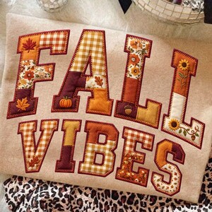 Op de afbeelding: Een beige sweatshirt met de tekst FALL VIBES in grote blokletters. Elke letter is gevuld met een patchwork van herfstachtige stoffen, waaronder oranje gingham, bloemenpatronen en geborduurde motieven van pompoenen en bladeren.