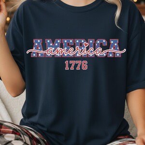 May include: Navy blue t-shirt with the word "AMERICA" in red and blue, decorated with white stars. The word "America" is written in cursive below the word "AMERICA". The number "1776" is printed in red below the word "America".