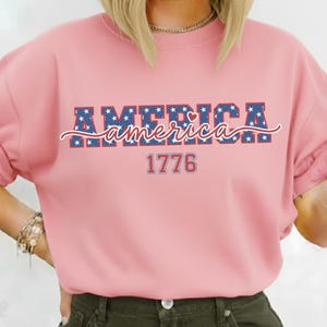 May include: A light pink sweatshirt with the word "AMERICA" in blue letters filled with white stars. The word "america" is written in cursive below, and the number "1776" is below that. The sweatshirt is a patriotic design.