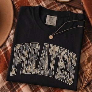 May include: A black short-sleeved t-shirt featuring the word PIRATES in large, arched, block-style letters with a camouflage pattern fill. The shirt has a crew neckline and a label at the inner collar that reads COMFORT COLORS S P CH.