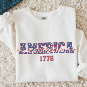May include: A white sweatshirt with the word "AMERICA" in blue letters filled with white stars, and the word "america" in a cursive font. The number "1776" is below the word "america". The sweatshirt has long sleeves.