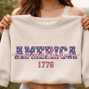 May include: A cream-colored sweatshirt with the word "America" in a patriotic design. The word "America" is in blue block letters with white stars, and a red heart. The year "1776" is below in red.