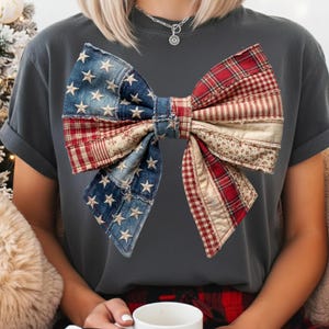 May include: A dark grey t-shirt with a large bow design. The bow is a patchwork of denim with white stars, red plaid, and floral fabric. The shirt has short sleeves and a crew neck.