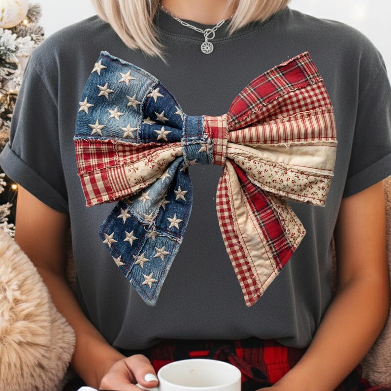 May include: A dark grey t-shirt with a large bow design. The bow is a patchwork of denim with white stars, red plaid, and floral fabric. The shirt has short sleeves and a crew neck.