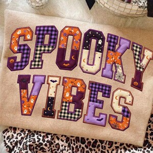 May include: A beige sweatshirt featuring the text SPOOKY VIBES in large, fabric-appliqued letters. Each letter is constructed from various patterned fabrics including orange pumpkins, white ghosts, black bats, spiderwebs, and purple plaid.