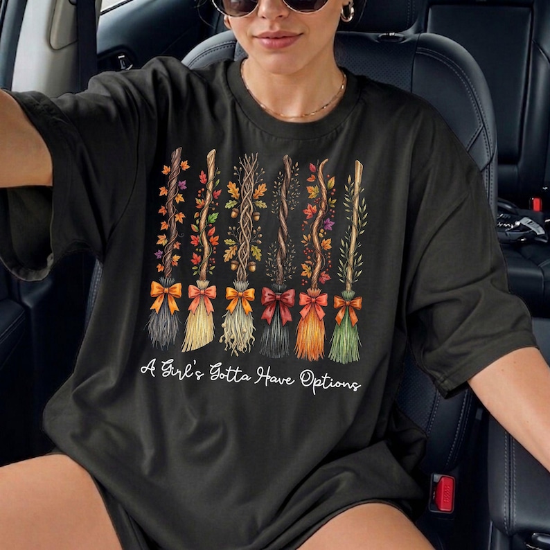 May include: Black t-shirt featuring a design of six decorative brooms with colorful bows and autumn foliage. The text "A Girl's Gotta Have Options" is printed below the brooms. The brooms have different colors and textures.
