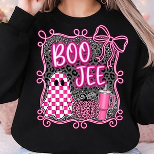 May include: A black sweatshirt featuring a graphic print with a pink neon-style border and a large pink bow. The design includes the text "BOO JEE" in pink, a pink and white checkered ghost, a pink leopard print pumpkin, and a pink tumbler cup.