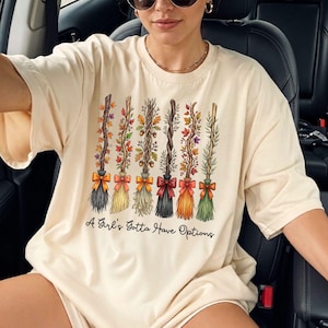 May include: Cream-colored t-shirt featuring a design of six decorative broomsticks with colorful autumn foliage and bows. The text "A Girl's Gotta Have Options" is printed below the broomsticks.