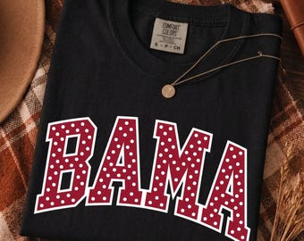 May include: A black short-sleeved t-shirt featuring the word BAMA in large, arched, maroon block letters with a white polka dot pattern. The shirt has a crew neckline and a label at the inner collar that reads Comfort Colors size S.