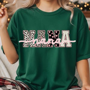 May include: A dark green t-shirt with the word "NANA" in a decorative font. The letters have a pink outline and are filled with leopard print, hearts, and polka dots. A cursive "nana" is written underneath.