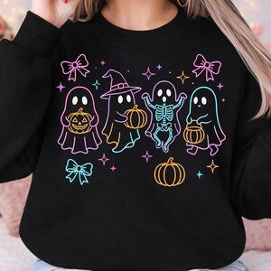 May include: A black long-sleeve sweatshirt featuring a neon-style graphic print of four ghosts, one wearing a witch hat and one with a visible skeleton ribcage, holding orange pumpkins. The design includes pink bows and colorful four-pointed stars.