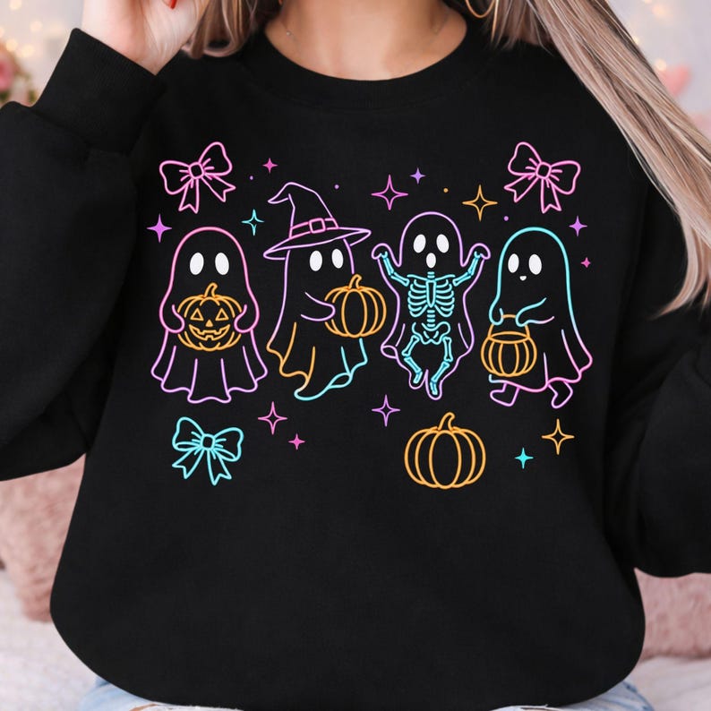 May include: A black long-sleeve sweatshirt featuring a neon-style graphic print of four ghosts, one wearing a witch hat and one with a visible skeleton ribcage, holding orange pumpkins. The design includes pink bows and colorful four-pointed stars.