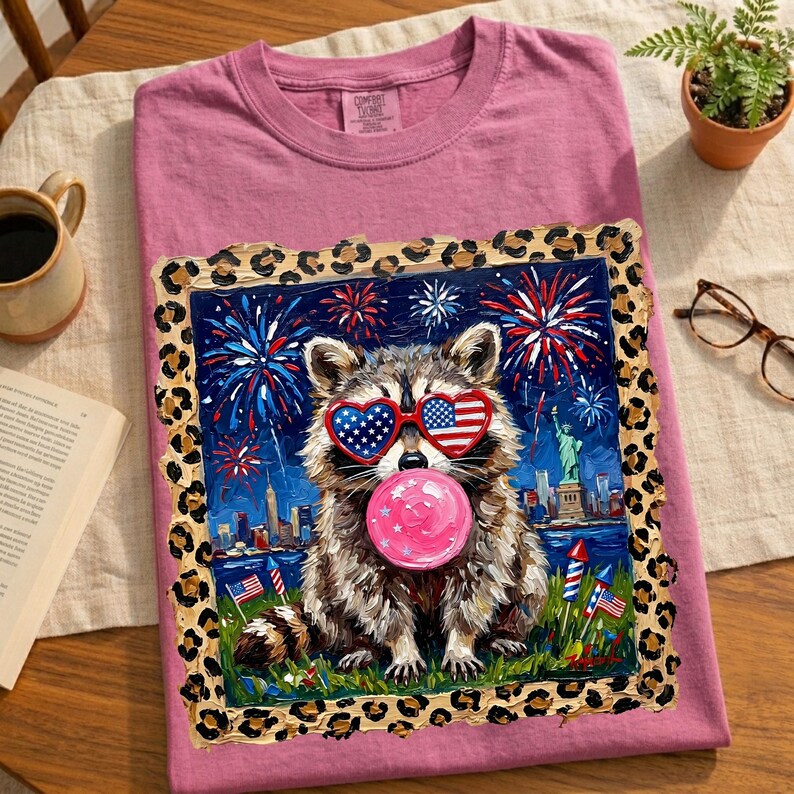 May include: A pink t-shirt featuring a raccoon wearing heart-shaped sunglasses with American flag designs, blowing a pink bubble. The graphic includes fireworks, the Statue of Liberty, and a cityscape. The design is framed with a leopard print border.