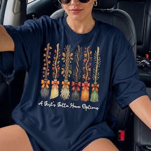 May include: Navy blue t-shirt featuring a design of decorative brooms with autumn leaves and bows. The text "A Girl's Gotta Have Options" is printed below the brooms. The shirt is a crew neck style.