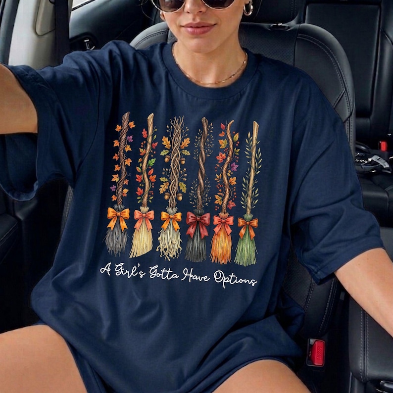 May include: Navy blue t-shirt featuring a design of decorative brooms with autumn leaves and bows. The text "A Girl's Gotta Have Options" is printed below the brooms. The shirt is a crew neck style.
