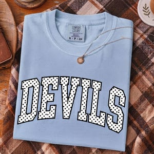 May include: A light blue t-shirt featuring the word DEVILS in a large, arched, black-and-white polka dot font across the chest. The shirt has a Comfort Colors brand label at the inner neckline and is displayed with a thin gold-toned necklace.